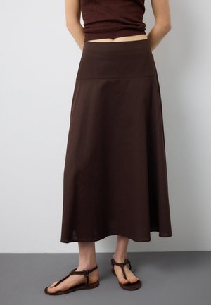 Donna che indossa una gonna marrone scuro a vita alta, lunga fino alla caviglia, abbinata a sandali bassi marroni, in piedi contro un muro grigio.