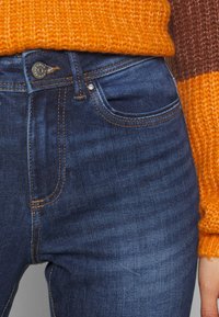 Mörkblå denimjeans med fem fickor, synlig sömnad och knäppning med knapp. Åtföljs av en texturerad orange stickad tröja.