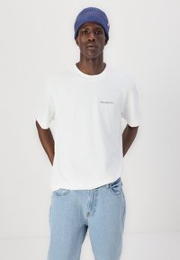 Tricou alb din bumbac, cu mâneci scurte și guler rotund, având un mic logo negru pe piept. Purtați cu blugi din denim de culoare albastră deschisă și o căciulă albastră cu dungi.
