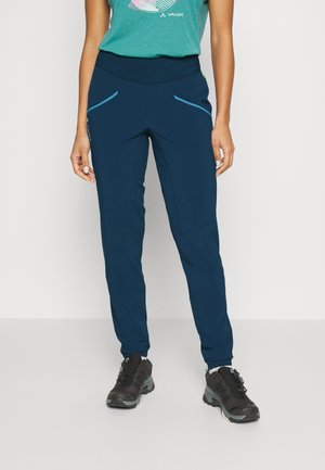Kobieta ubrana w ciemnoniebieskie spodnie sportowe z jasnoniebieskimi akcentami kieszeni i czarne buty trekkingowe stoi na jednolitym tle.