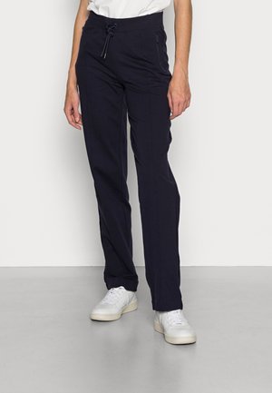 Persoon die zwarte broek met trekkoord en witte sneakers draagt, staand tegen een effen lichtgrijze achtergrond.