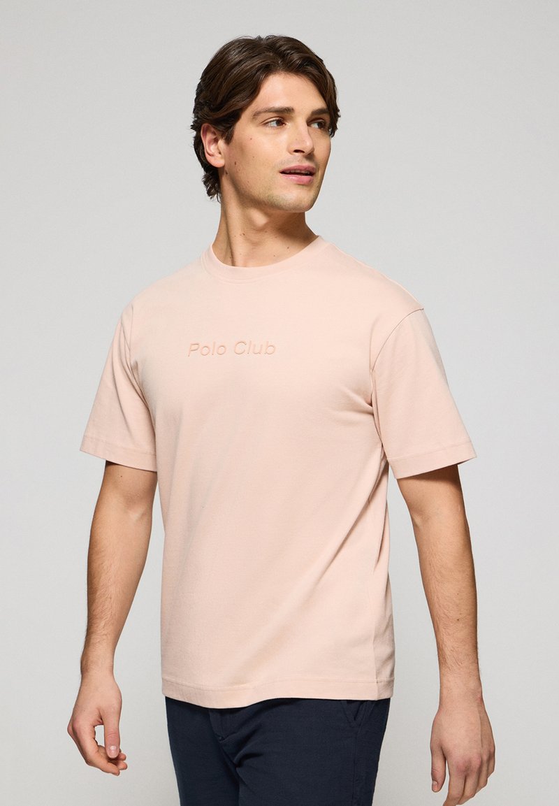 Camiseta de manga corta rosa pálido con cuello redondo que presenta un logo en relieve de "Polo Club" en la parte frontal. Material de algodón suave.
