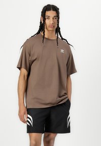 Brun bomulds t-shirt med rund hals og korte ærmer, med et lille hvidt Adidas-logo på brystet. Sammen med sorte shorts.