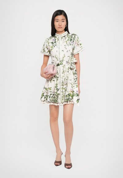Bloemen wikkeljurk met een witte basis en groene en paarse bloemenpatronen, korte mouwen, knopen en een aansnoer taille; geaccessorieerd met een roze clutch.