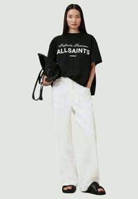 T-shirt de algodão preto com texto branco, combinado com calças largas brancas. Os acessórios incluem uma bolsa preta e sandálias pretas rasas.