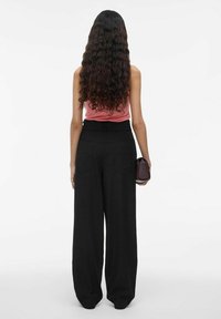 Zwarte, wijde broek met achterzakken, gecombineerd met een roze mouwloos top. Het model houdt een kleine, rechthoekige bordeauxrode tas vast.