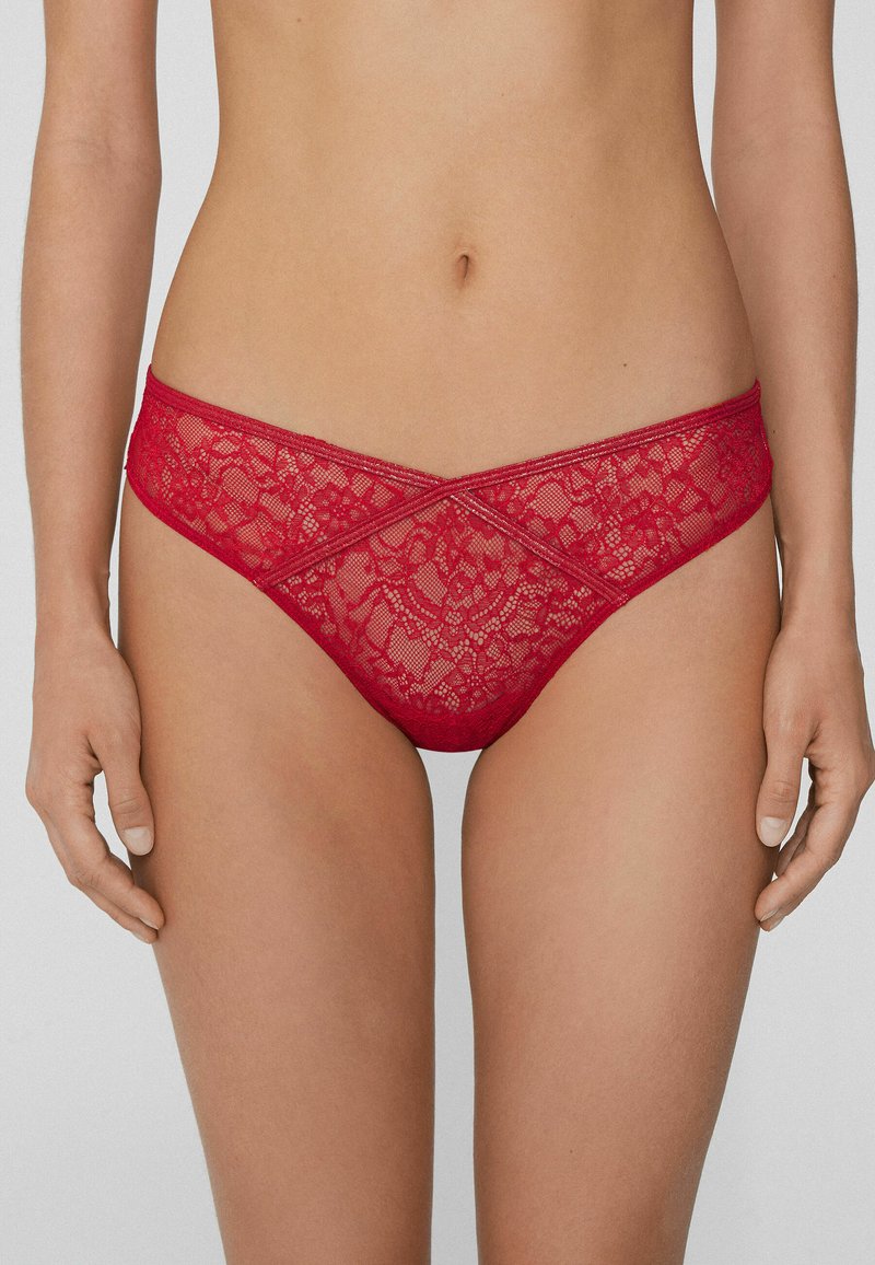 Culotte bikini en dentelle rouge avec un motif floral, une forme triangulaire, et une ceinture délicate ornée d'un détail en croix à l'avant.