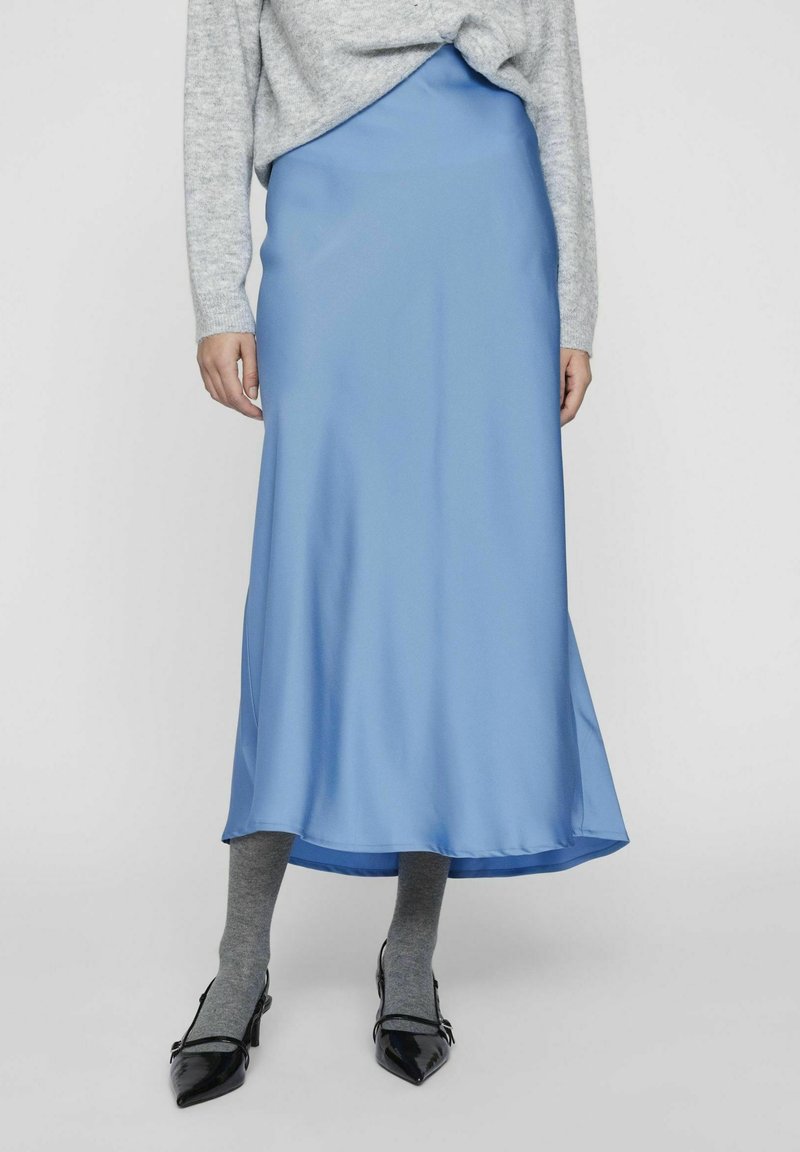Femme portant une jupe midi en satin bleu clair, un pull gris, des collants gris et des escarpins en cuir verni noir, debout devant un arrière-plan gris.