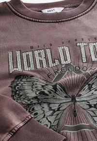 Sweat-shirt mauve texturé avec des embellissements métalliques. Présente un graphisme de papillon et le texte "WORLD TOUR" en clous argentés. Poignets côtelés.