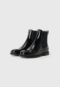 Une paire de bottines Chelsea en cuir noir brillant avec des panneaux élastiques sur les côtés et des talons bas sur un fond blanc.