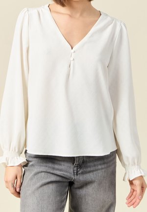 Femme portant un chemisier blanc à manches longues avec un col en V et deux boutons, associé à un jean denim gris.