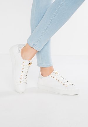 Osoba ubrana w jasnoniebieskie dżinsy i białe niskie trampki z złotymi przelotkami i teksturowanym detalem na pięcie, stojąca na białej powierzchni.