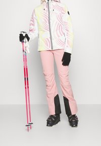 Ski bunda v bílé barvě s růžovými a žlutými vlnovkami, doplněná o růžové kalhoty s černými akcenty. Černé rukavice a lyžařské hole dotvářejí celkový look.