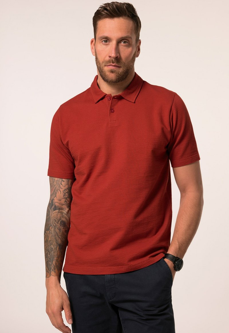 Röd polo med ett strukturerat mönster, korta ärmar och en krage med tre knappar. Bärs med mörka byxor och en svart armbandsklocka.