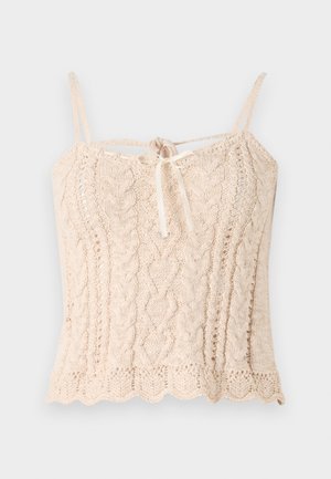 Camisola de malha em bege claro com uma textura canelada, apresentando alças finas e um detalhe em laço atado na frente. Bainha com bordas onduladas.