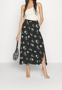 Jupe maxi noire à fleurs avec fentes latérales, ornée de feuilles vertes et de fleurs roses. Associée à un haut blanc asymétrique et des talons transparents.