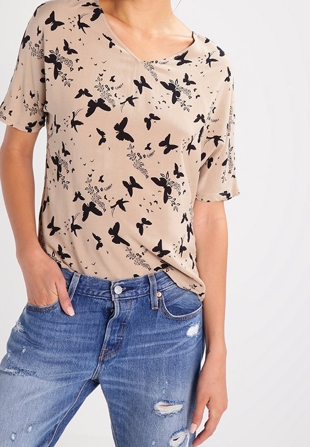 Blusa beige de manga corta con escote en V, con un patrón de mariposas negras y flores. Combinada con jeans de denim azul desgastados.