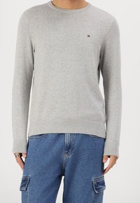Personne portant un pull ras du cou gris clair avec un petit logo sur la poitrine et un pantalon cargo en denim bleu ample avec des poches latérales.