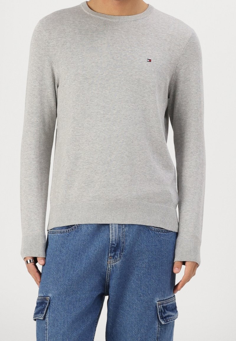 Personne portant un pull ras du cou gris clair avec un petit logo sur la poitrine et un pantalon cargo en denim bleu ample avec des poches latérales.