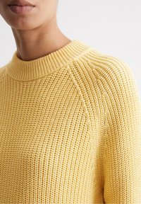 Primo piano di una persona che indossa un maglione lavorato a maglia gialla con un colletto alto a coste.