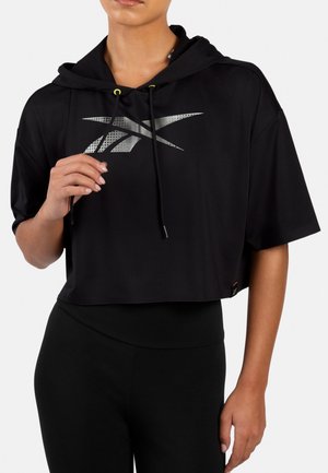 Person trägt schwarzen kurzen Hoodie mit silbernem Logo und schwarzen Leggings, hält eine Kordel nahe der Brust.