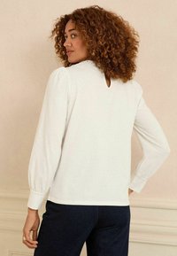 Blusa bianca a maniche lunghe con scollatura rotonda e dettagli in pizzo. Apertura a occhio di chiave sul retro e polsini arricciati. Tessuto morbido e texture.