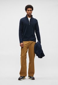 Ναυτική ζακέτα με φερμουάρ, καφέ cargo παντελόνια και multicolored αθλητικά παπούτσια. Το μοντέλο κρατά μια ναυτική ζακέτα, στέκεται μπροστά σε ανοιχτό γκρι φόντο.