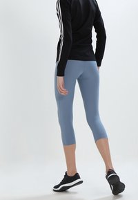 Blå capri leggings lavet af strækbart materiale, parret med en sort langærmet top med hvide striber langs ærmerne og sorte sportsko.
