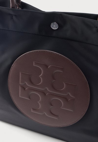 Sac fourre-tout en tissu noir avec une pièce circulaire en cuir marron présentant un logo stylisé et symétrique, ainsi qu'une fermeture à bouton-pression en métal.