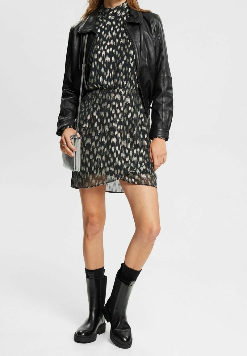 Femme portant une robe à motifs noir et blanc, une veste en cuir noire, des bottes noires mi-mollet et tenant un petit sac bandoulière argenté.