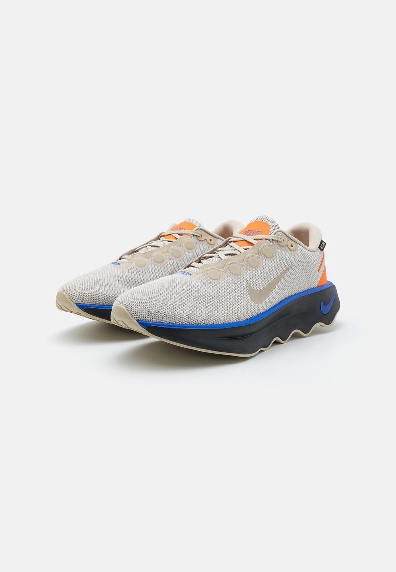 Zapatillas deportivas de malla beige con acentos naranja y azul, que presentan una suela negra curva y un prominente logo de Nike en blanco en el lado.