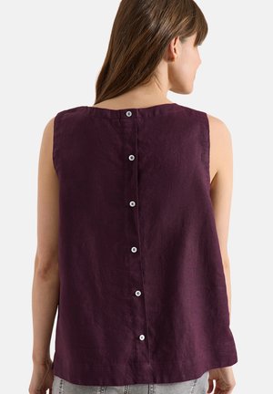 Frau trägt ein ärmelloses dunkelviolettes Top mit weißen Knöpfen auf dem Rücken und steht vor einem einfachen hellen Hintergrund.