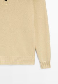 Maglione beige a maglia con colletto, tre bottoni neri, polsini e orlo a costine, caratterizzato da una texture morbida e una vestibilità rilassata.