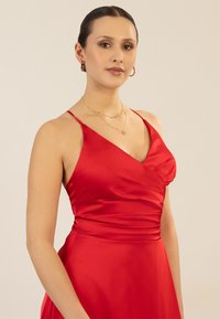 Rotes Satinabendkleid mit drapierter Brustpartie und dünnen Trägern. Verfügt über eine taillierte Taille und einen fließenden Rock, ergänzt durch goldene Schmuckakzente.