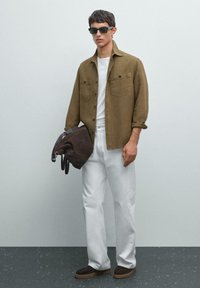 Braune Button-Down-Hemd mit zwei Brusttaschen, weißes T-Shirt, weiße Hose, braune Wildlederschuhe und eine braune Tasche mit einem Gurt.