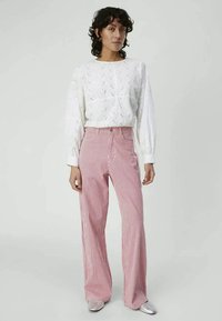 Γυναίκα με σγουρά σκούρα μαλλιά, φορώντας μια άσπρη υφασμάτινη μπλούζα μακρυμάνικη και παντελόνι με ψηλή μέση σε ροζ και άσπρες ρίγες με φαρδιά ποδιά.