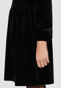 Schwarzes Samtkleid mit langen Ärmeln, gerafftem Taillenbereich und glatter Textur. Der Saum ist gerade, und die Ärmelsäume sind elastisch.