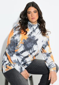 Frau mit langen dunklen Haaren, die einen Pullover mit abstraktem Muster in Weiß, Orange und Grau sowie dunkelgraue Jeans trägt, sitzt auf einem Hocker vor einem weißen Hintergrund.