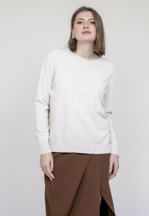 Jeune femme aux cheveux mi-longs porte un pull blanc à manches longues et une jupe marron fendue à l'avant, debout devant un fond uni.