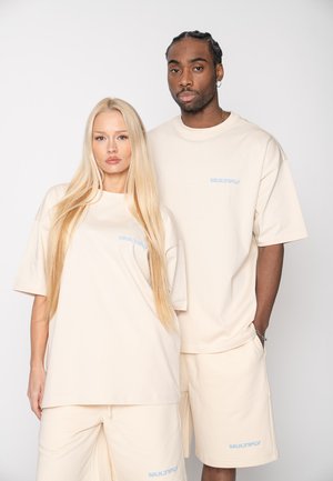 Man and woman standing, wearing matching oversized beige t-shirts and shorts with "MULTIPLY" logo in light blue.