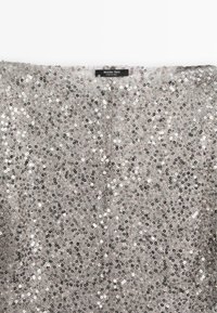 Tissu recouvert de sequins argentés avec une étiquette visible « Massimo Dutti » cousue en haut au centre, présentant des décorations denses et réfléchissantes.