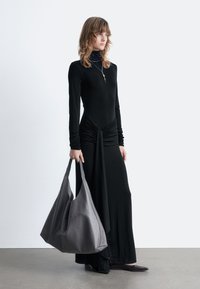 Langes schwarzes Kleid mit geraffter Taille, langen Ärmeln und leichtem Glanz. Graue Leder-Hobo-Tasche mit geschwungenen Nähten und glatter Textur.