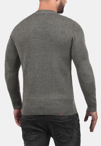 Grå langærmet sweater med ribbet kant og manchetter, rund halsudskæring, og med albuepatches. Stoffet ser blødt ud med en glat tekstur.