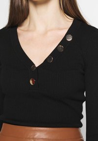 Pull noir côtelé avec un col en V et cinq boutons brillants sur une épaule, associé à une jupe en cuir marron lisse.