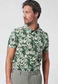 Camicetta verde a maniche corte con un motivo a foglie crema, colletto a camicia e vestibilità slim. Indossata con pantaloni grigi, caratterizzati da una vita standard.