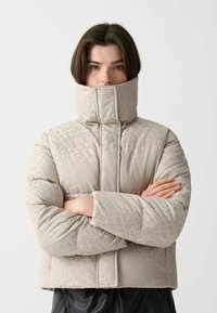 Vadderad jacka i ljusbeige med subtilt leopardmönster. Hög krage, kviltad design och vida ärmar. Framslutning med dolda knappar.