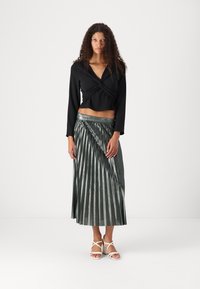 Black long-sleeve cropped top with twisted front detail, paired with a metallic green pleated maxi skirt. Sandals feature white straps.