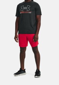 Czarny T-shirt z krótkim rękawem z dużym białym i czerwonym logo Under Armour, w zestawie z czerwonymi szortami i czarnymi butami sportowymi.