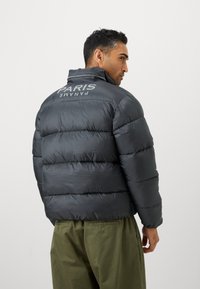 Tamno siva puffer jakna s horizontalnim prošivanjem, visokim ovratnikom i reflektirajućim logotipom "PARIS PANAME" na leđima. Tekstuirana tkanina stvara dojam topline.
