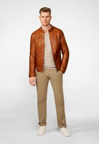 Brun læderjakke med en rund krave, beige sweater under, og khaki bukser. Modellen har hvide sneakers på og står foran en lys baggrund.